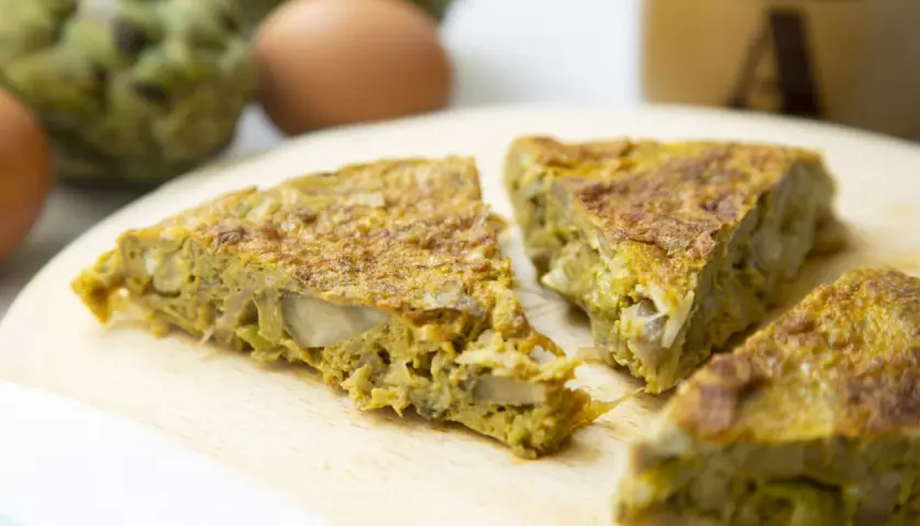Fette di frittata con pecorino e verdure su tagliere di legno, con uova e carciofi sullo sfondo.