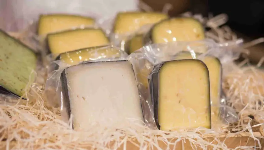 Vari tipi di formaggio fuori dal frigo, confezionati singolarmente e disposti su paglia in una cesta di legno.