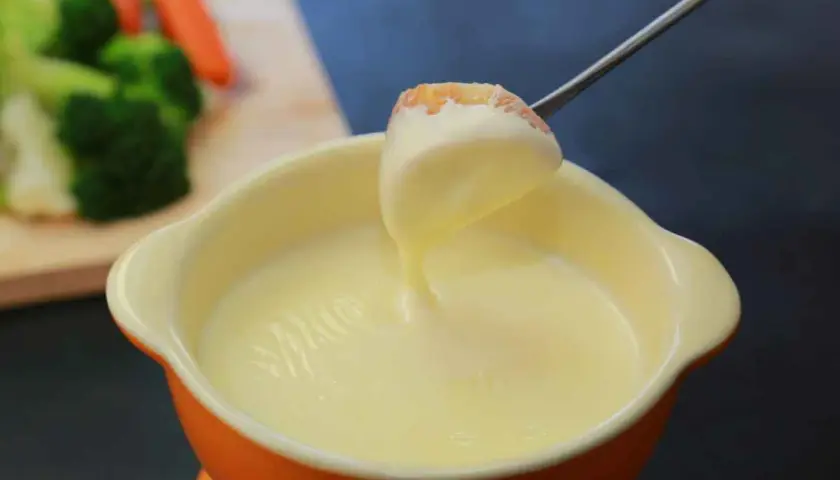 Una forchetta intinge un pezzo di pane in una cremosa fonduta per pasta. Sullo sfondo, verdure fresche.