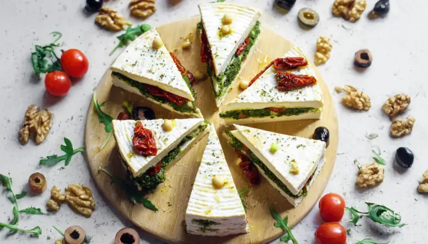 Fette di primo sale al forno, farcite con pesto e pomodori secchi, disposte a raggiera su un tagliere, con noci e pomodorini intorno.