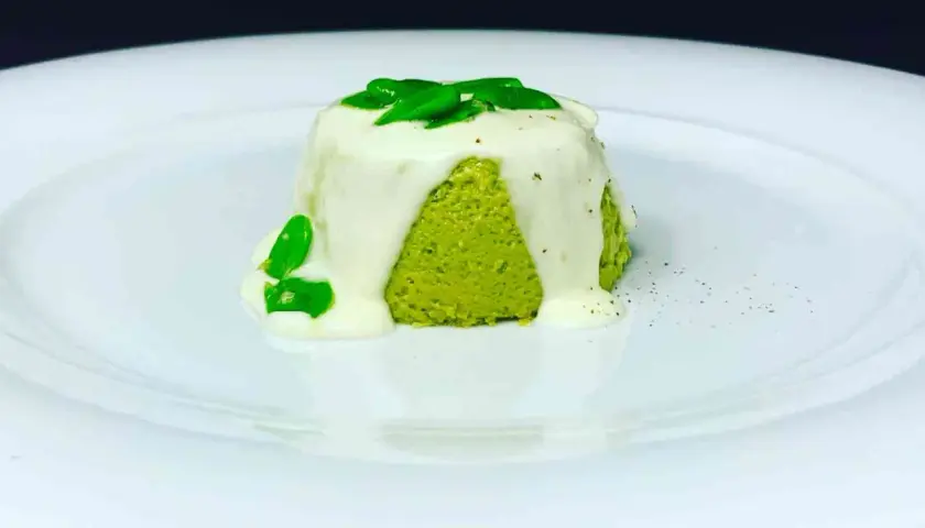 Sformato di pecorino verde brillante, ricoperto da una salsa bianca cremosa e guarnito con foglie verdi fresche.