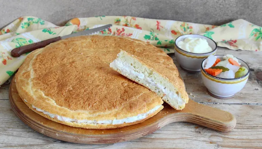 Torta salata con formaggi avanzati, soffice e dorata, con una fetta tagliata che rivela il ripieno cremoso. Accanto, ciotoline con salse e verdure sottaceto.