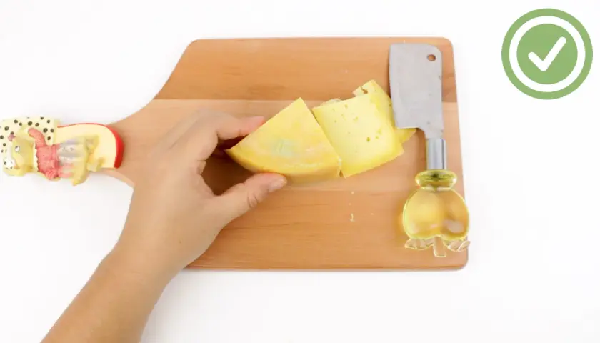 Mano che tiene un pezzo di formaggio giallo su un tagliere di legno, pronto per imparare come tagliare il formaggio a spicchi con un coltello.