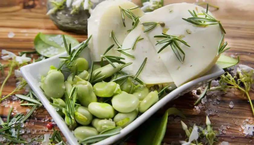 Baccelli freschi e fette di pecorino guarnite con rosmarino su un tagliere di legno.