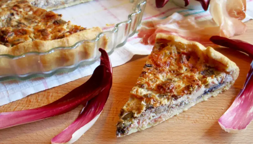 Una fetta di torta salata con taleggio, ricca e dorata, accanto a foglie di radicchio e prosciutto crudo.