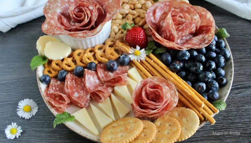 Presentazione tagliere aperitivo con salumi a rosa, formaggi, mirtilli, fragole, pretzel e grissini.