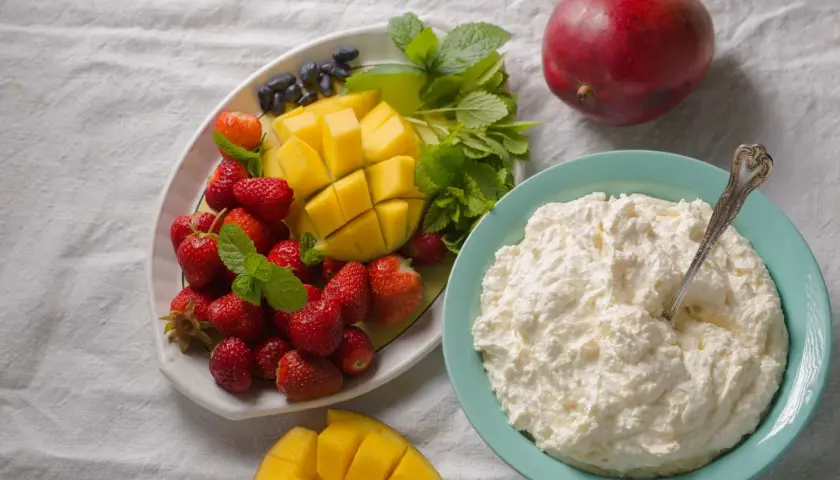 Un piatto di fragole, mango e mirtilli accanto a una ciotola di cosa sono i fiocchi di latte, con un mango intero sullo sfondo.