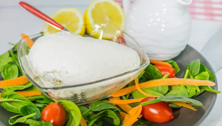 Un fresco quark formaggio in una ciotola, circondato da spinaci, carote e pomodorini. Limoni e una brocca bianca sullo sfondo.