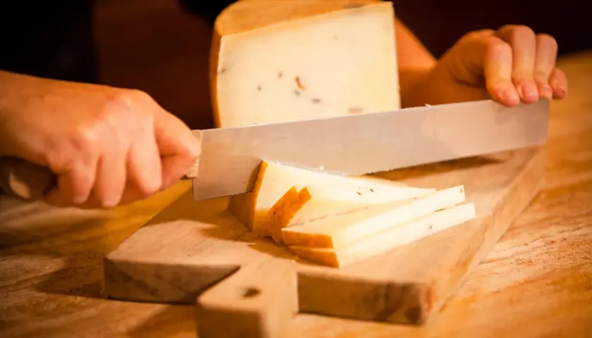 Mani che tagliano formaggio su un tagliere di legno. Fette sottili e un pezzo più grande di formaggio stagionato.
