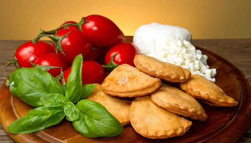Panzerotti pugliesi dorati, pomodorini freschi, mozzarella a cubetti e basilico profumato su un tagliere di legno.