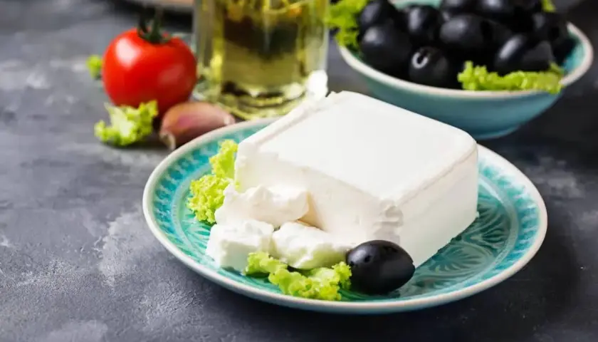 Un blocco di feta su un piatto turchese, con olive, pomodoro e olio d'oliva. La feta proprietà è l'ingrediente principale di questa deliziosa insalata greca.