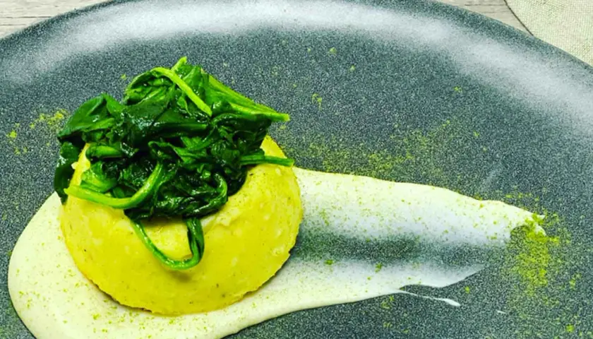 Flan di pecorino giallo, sormontato da spinaci freschi, su una crema bianca e polvere verde su piatto scuro.