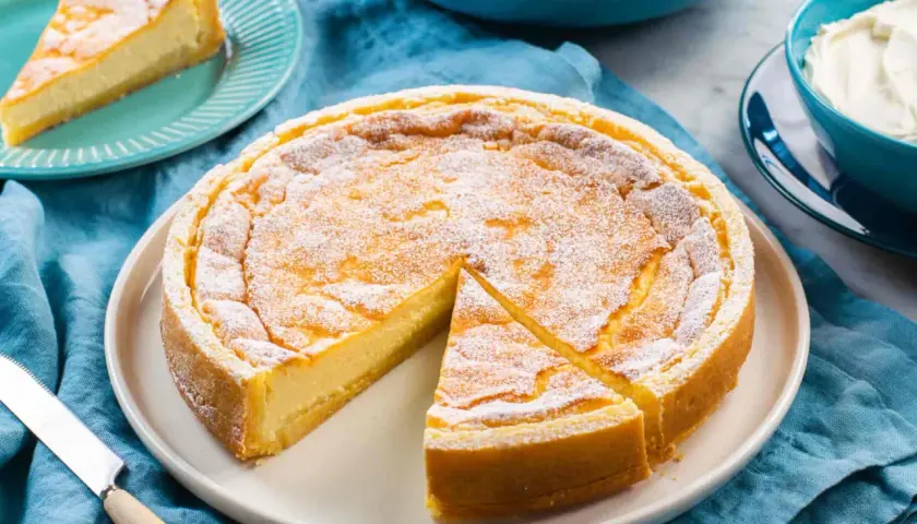 Torta soffice e dorata, simile a un quark formaggio, cosparsa di zucchero a velo. Una fetta è già servita.