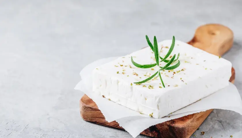 Un blocco di feta su un tagliere di legno, guarnito con rosmarino e spezie. Ricorda che la feta contiene lattosio.