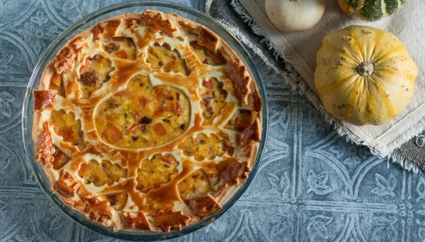 Torta salata zucca salsiccia e stracchino, con decorazione a intreccio e ripieno dorato. Accanto, zucche decorative.