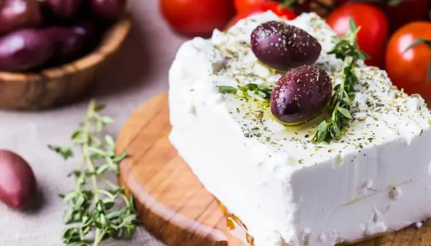 Un blocco di feta greca condita con erbe aromatiche e olive su un tagliere di legno, con pomodori e olive in sottofondo.
