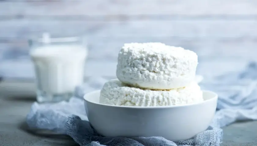 Due forme di ricotta fresca in una ciotola bianca, con un bicchiere di latte sullo sfondo. La ricotta si può congelare per conservarla più a lungo.
