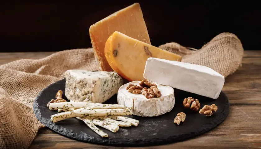 Selezione di formaggi grassi: un pezzo di formaggio erborinato, un brie, un formaggio stagionato e bastoncini di formaggio alle erbe, con noci e tela di juta su un tagliere di ardesia.