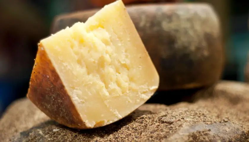 Un pezzo di formaggio sardo stagionato, con crosta scura e pasta gialla, riposa su un letto di spezie macinate.