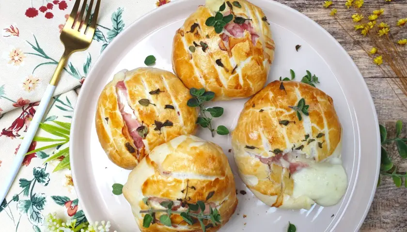 Quattro fagottini dorati con prosciutto e **tomino fuso** che cola, guarniti con erbe aromatiche su un piatto rosa.