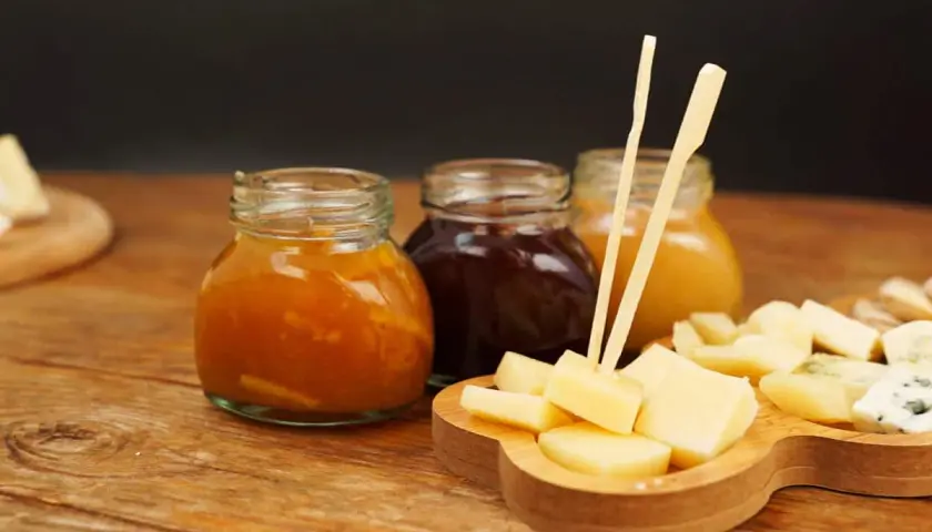 Tre vasetti di marmellata, uno arancione, uno scuro e uno giallo, accompagnano un tagliere di formaggio a cubetti, pronto per essere gustato.