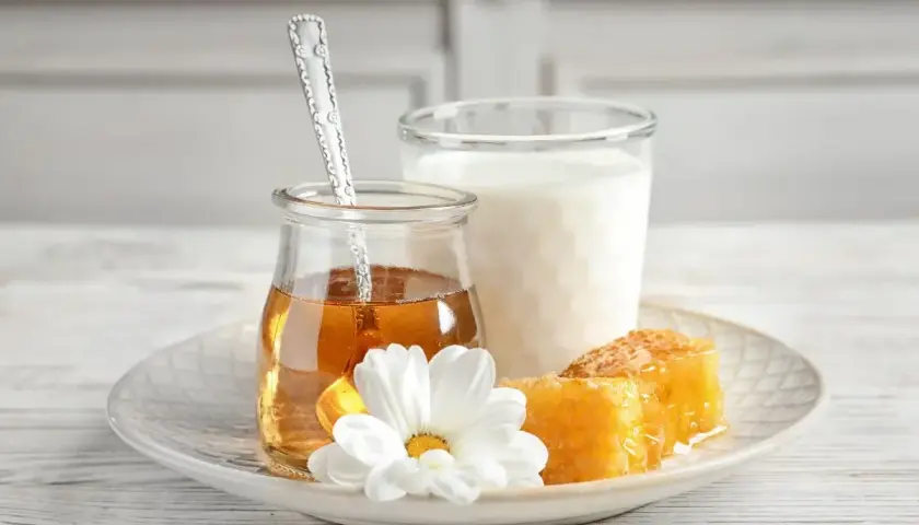 Un bicchiere di latte, un vasetto di miele dorato con un cucchiaino, e favi di miele su un piatto con un fiore bianco. Alcuni credono che latte caldo e miele faccia male.
