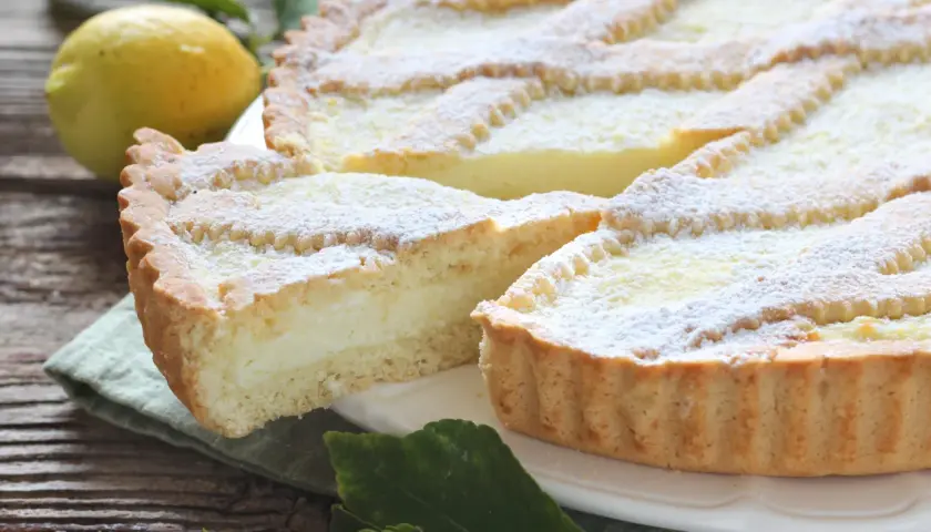 Una fetta di crostata di ricotta, cosparsa di zucchero a velo, accanto a un limone e foglie verdi su un tavolo di legno.