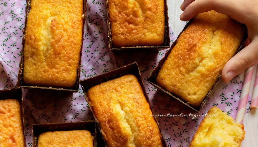 Plumcake yogurt dorati e soffici, pronti per essere gustati. Una mano ne afferra uno, mentre un pezzetto mostra la morbidezza interna.