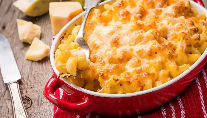 Mac and cheese dorato e filante in una teglia rossa, con pezzi di formaggio e una forchetta pronta a servirlo.