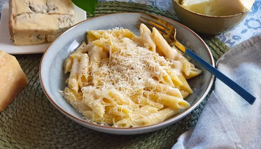 Piatto di penne cremose, condite con una ricca salsa di spaghetti ai 4 formaggi, guarnite con parmigiano grattugiato. Accanto, pezzi di formaggio fresco e stagionato.