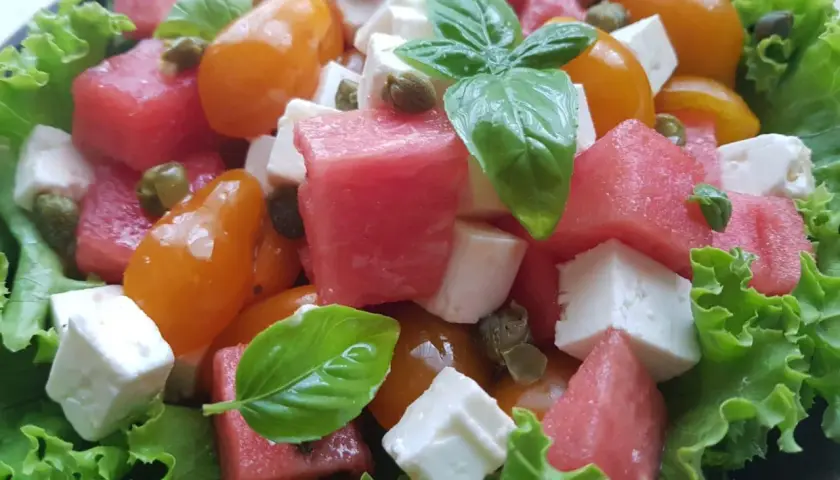 Insalata estiva con cubetti di formaggio di capra, anguria, pomodorini gialli, capperi e basilico fresco su letto di lattuga croccante.