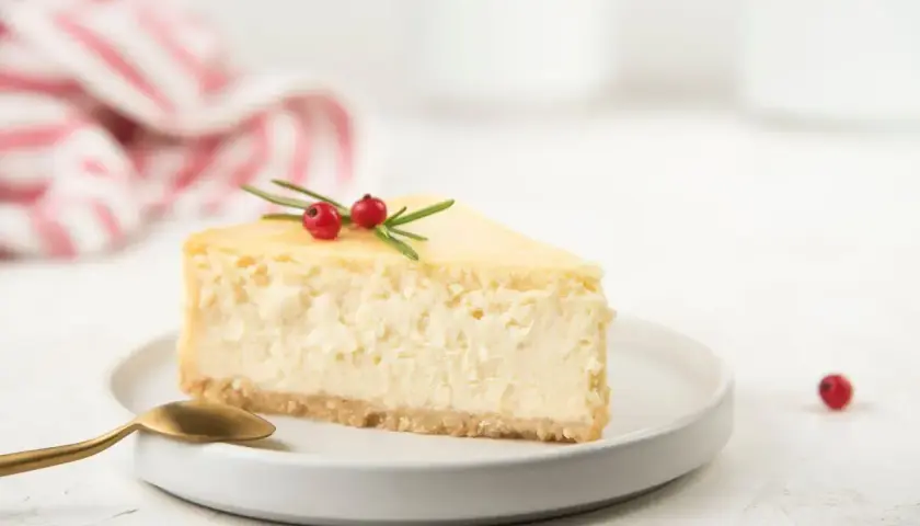 Una fetta di cheesecake con yogurt greco, guarnita con rosmarino e bacche rosse, su un piatto bianco con un cucchiaino dorato.