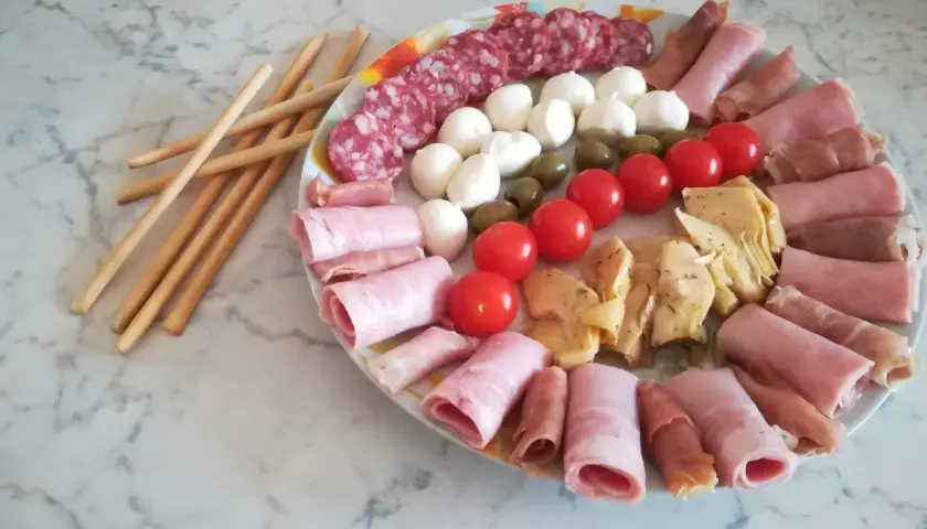 Un'idea per un tagliere di salumi: salame, mozzarelline, olive, pomodorini, carciofini e involtini di prosciutto cotto e crudo, con grissini a lato.