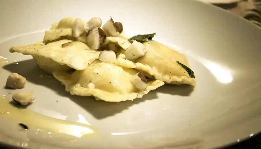 Ravioli pere e pecorino, conditi con nocciole tritate e una foglia di salvia, su un piatto bianco con un filo d'olio.