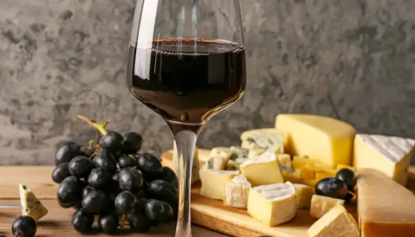 Un bicchiere di vino rosso accompagna un invitante tagliere di formaggi misti, con uva nera e un coltello da formaggio.