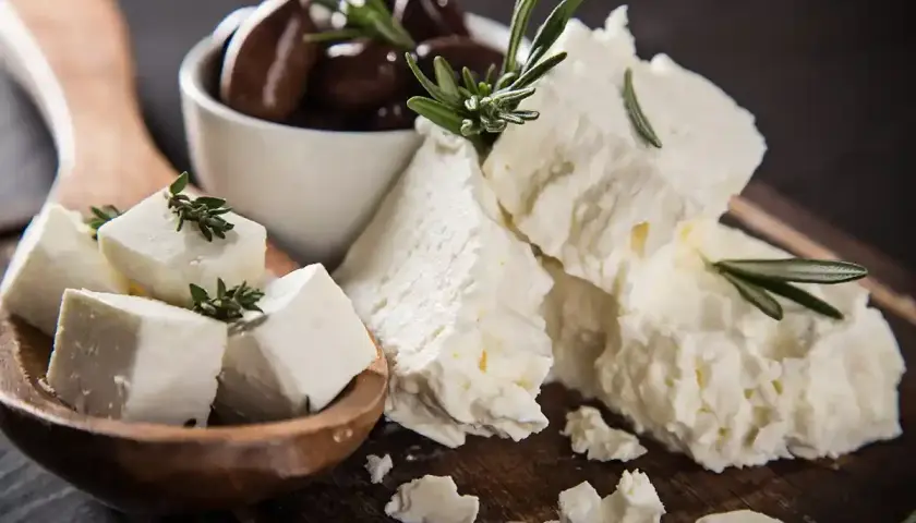 Feta, un formaggio ricco di proteine per 100g, servito con olive nere e rametti di rosmarino su un tagliere di legno.