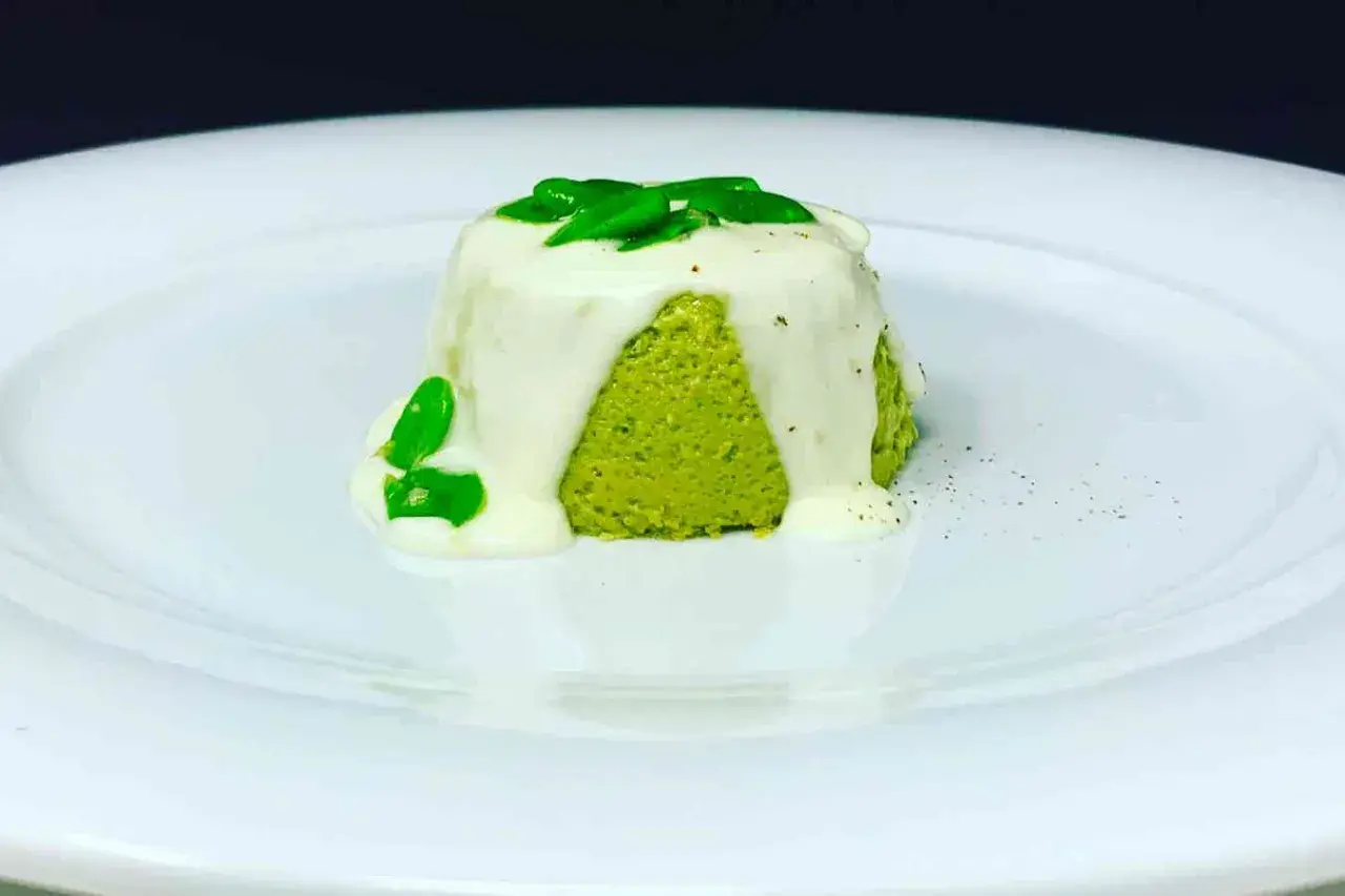 Sformato di pecorino verde brillante, ricoperto da una salsa bianca cremosa e guarnito con foglie verdi fresche.