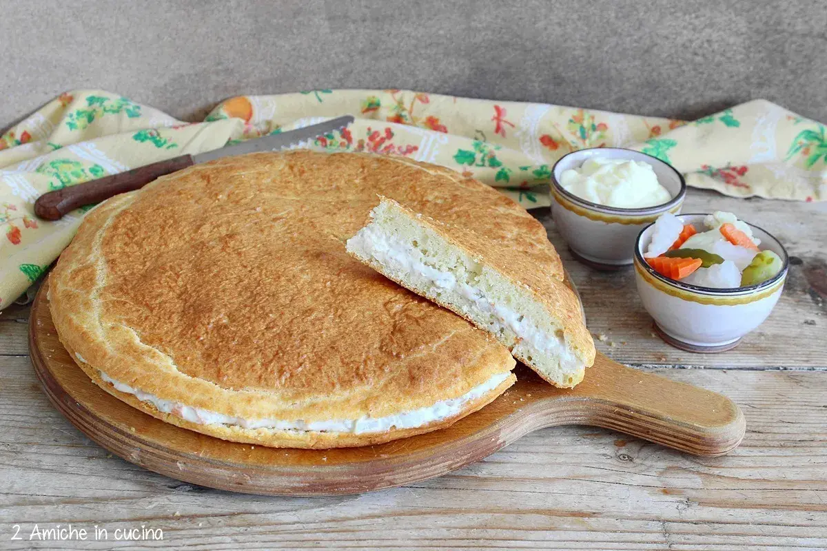 Torta salata con formaggi avanzati, soffice e dorata, con una fetta tagliata che rivela il ripieno cremoso. Accanto, ciotoline con salse e verdure sottaceto.