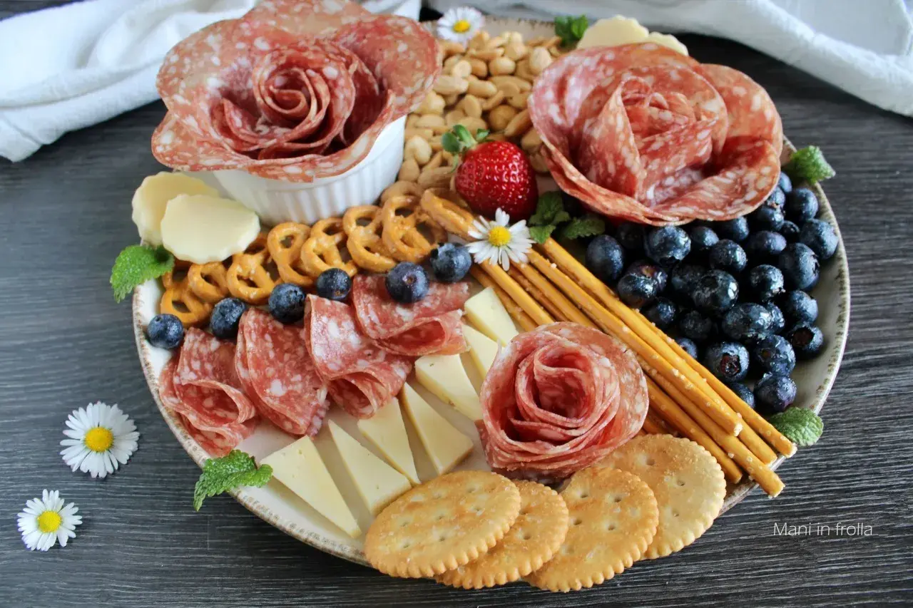 Presentazione tagliere aperitivo con salumi a rosa, formaggi, mirtilli, fragole, pretzel e grissini.