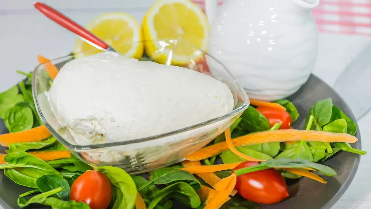 Un fresco quark formaggio in una ciotola, circondato da spinaci, carote e pomodorini. Limoni e una brocca bianca sullo sfondo.