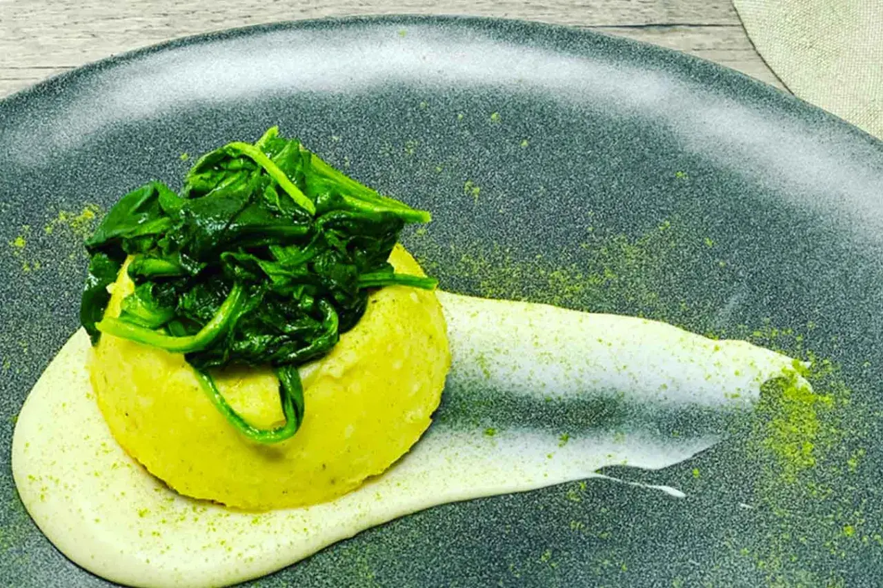Flan di pecorino giallo, sormontato da spinaci freschi, su una crema bianca e polvere verde su piatto scuro.