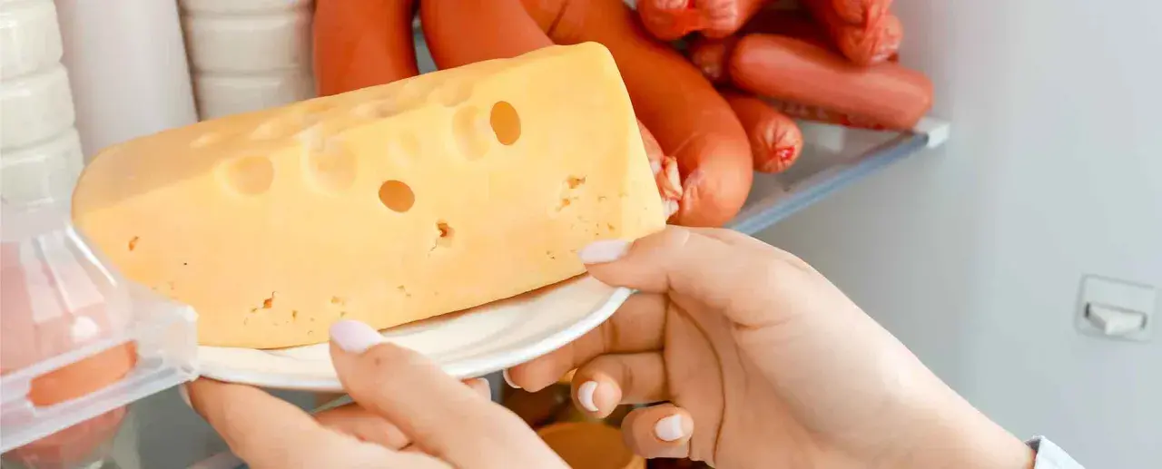 Mano prende un pezzo di formaggio svizzero da un frigorifero pieno di salumi. Formaggi salutari per uno spuntino.