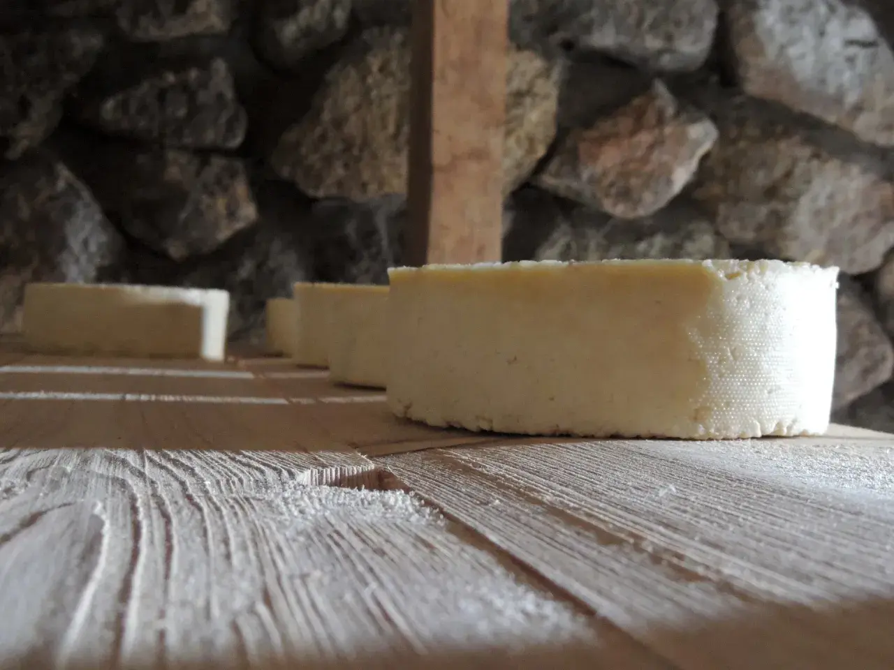 Formaggi freschi in fase di produzione, riposano su assi di legno grezzo. La pietra grezza fa da sfondo a questo momento di attesa.