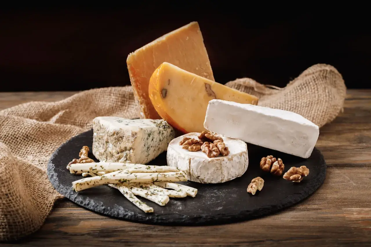 Selezione di formaggi grassi: un pezzo di formaggio erborinato, un brie, un formaggio stagionato e bastoncini di formaggio alle erbe, con noci e tela di juta su un tagliere di ardesia.