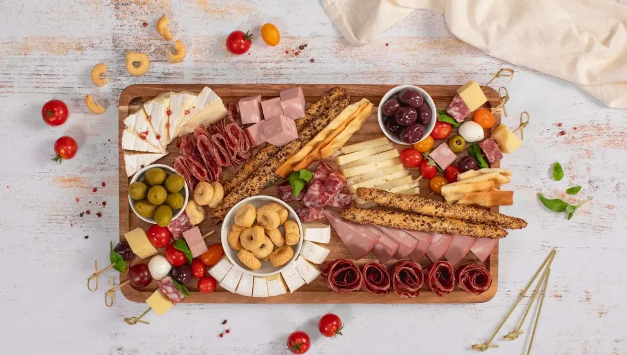 Un ricco antipasto salumi e formaggi su tagliere: salame, prosciutto, brie, taralli, olive e pomodorini.