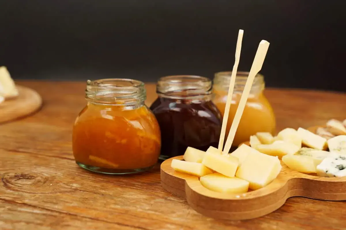 Tre vasetti di marmellata, uno arancione, uno scuro e uno giallo, accompagnano un tagliere di formaggio a cubetti, pronto per essere gustato.