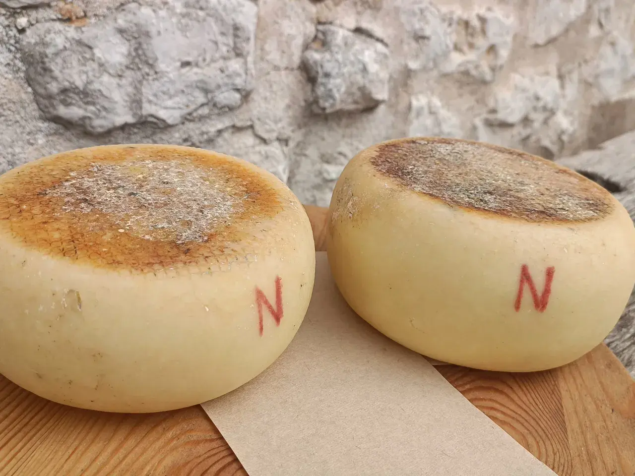 Due forme di pecorino semistagionato, con la crosta marcata e la lettera "N" rossa dipinta sul fianco, riposano su un tagliere di legno.