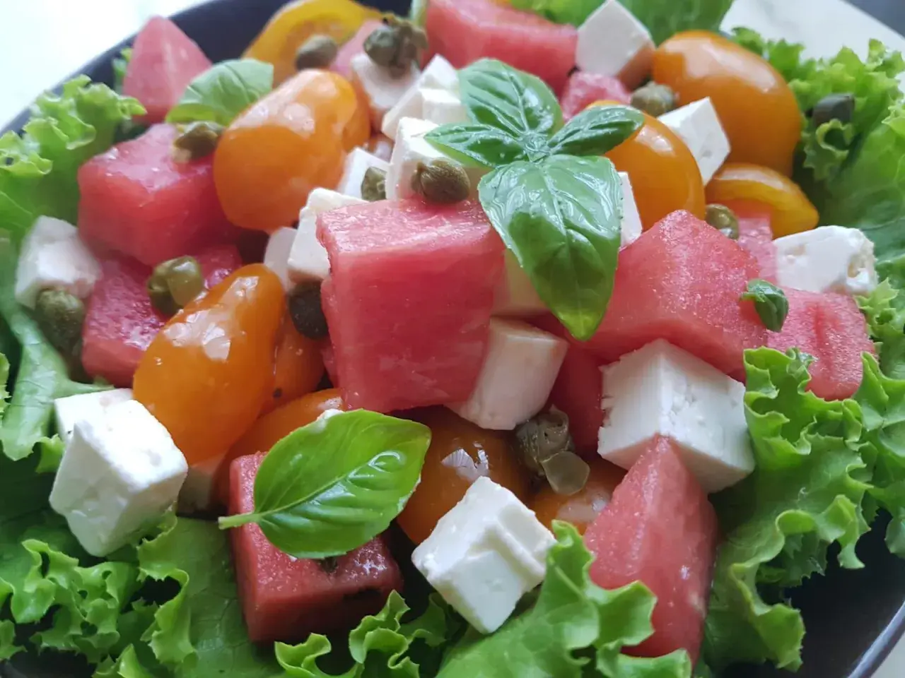 Insalata estiva con cubetti di formaggio di capra, anguria, pomodorini gialli, capperi e basilico fresco su letto di lattuga croccante.