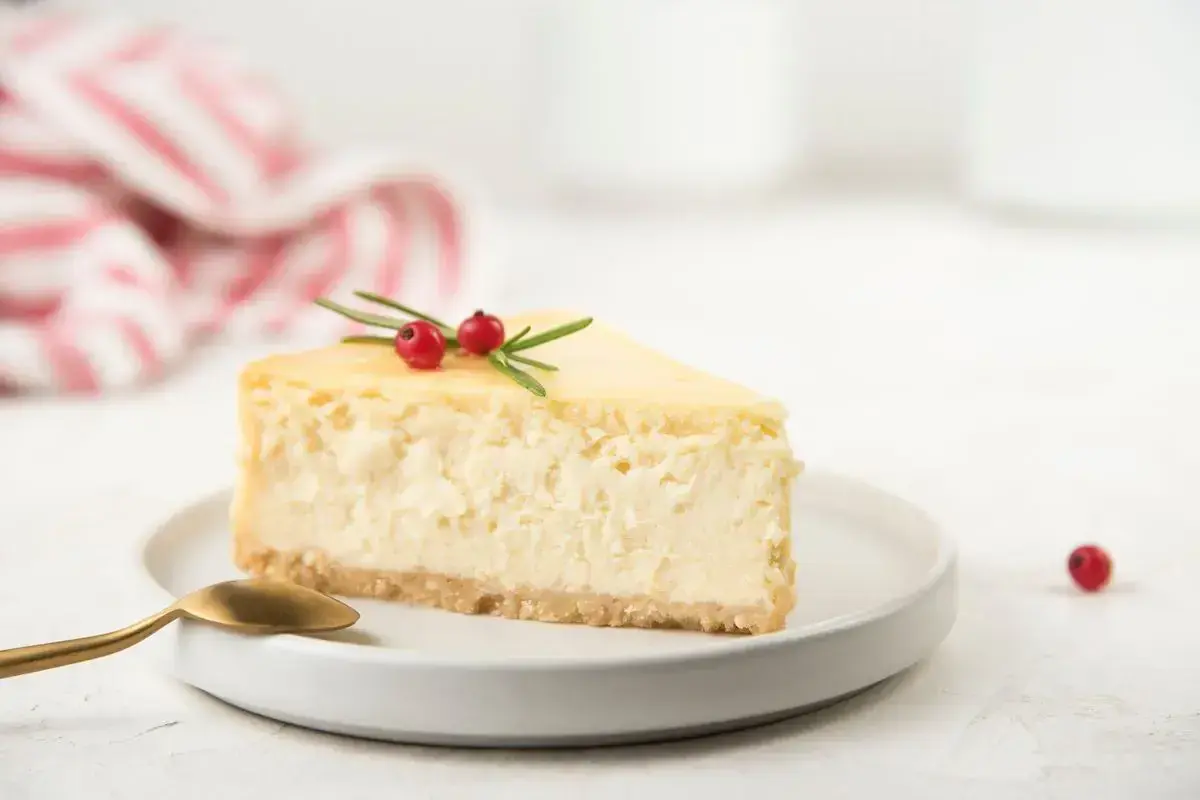Una fetta di cheesecake con yogurt greco, guarnita con rosmarino e bacche rosse, su un piatto bianco con un cucchiaino dorato.