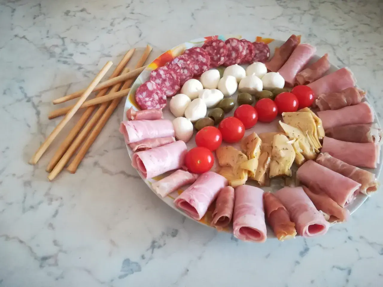 Un'idea per un tagliere di salumi: salame, mozzarelline, olive, pomodorini, carciofini e involtini di prosciutto cotto e crudo, con grissini a lato.